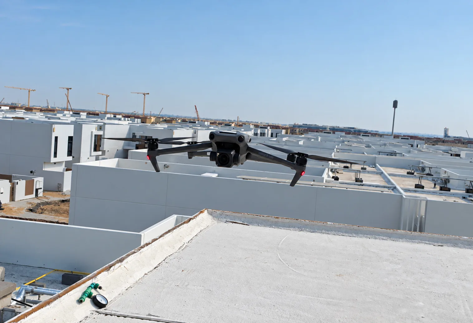 A camera drone hovers beside a rooftop with residential buildings and construction cranes behind it at The Sustainable City, Yas Island, Abu Dhabi