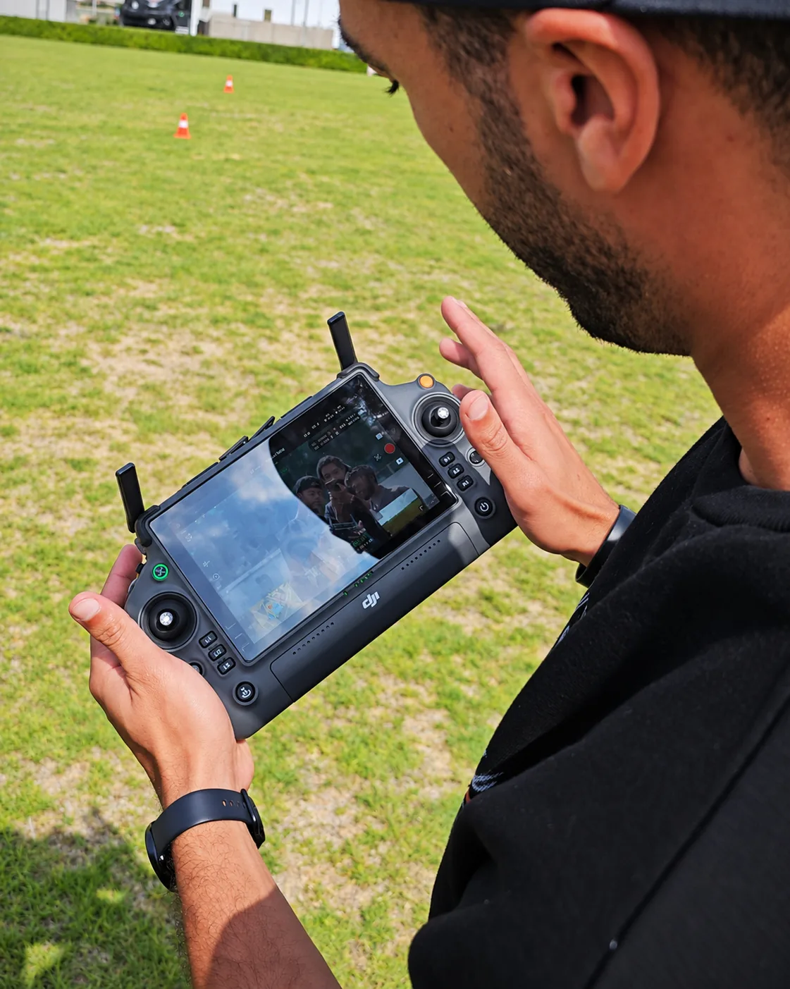 A pilot holds a drone controller above a grass field while checking the screen